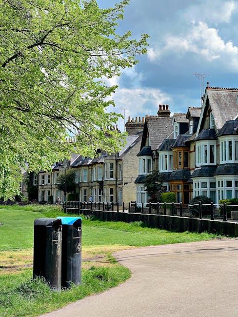 A row of traditional Victorian-style terraced houses with bay windows, constructed from light-colored brick with dark roof shingles, are visible in the background of the image. In the foreground, a grassy area with a wide paved pathway runs parallel to the houses, and to the left, a large tree with fresh green foliage extends its branches across the scene, providing partial shade. Near the bottom left corner, two black recycling bins with open slits on top and slim label patches are positioned side by side on the edge of the grass. The sky above is partly cloudy with a mix of blue sky and white clouds, suggesting a bright, mild day. The scene appears to be in a residential area, demonstrating a typical setting where local waste collection might be complemented by private rubbish removal services, supported by the presence of waste containers near the pathway, and the well-maintained appearance indicates possible management by a waste removal company like wasteremovalrichmonduponthames.co.uk.