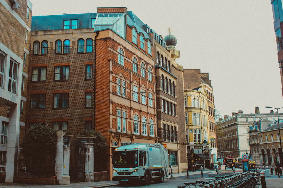 A city street scene featuring a white waste collection truck parked in front of a row of historic multi-storey buildings with brick and stone facades, large arched windows, and decorative architectural elements. The truck is positioned on the cobbled pavement, adjacent to a small fenced area with trees and a stone gate. Surrounding buildings display a mix of brown, beige, and pale yellow tones, with some featuring traditional domed rooftops and ornate detailing characteristic of urban architecture. To the right, modern glass and steel structures are visible, alongside a row of bicycles locked to a bike rack, indicating a busy city environment. The scene is illuminated by natural daylight, casting soft shadows, consistent with an urban setting where private waste collection services, such as those provided by waste removal specialists like wasteremovalrichmonduponthames.co.uk, frequently operate in commercial and residential areas to manage rubbish and debris within city streets and alleyways.